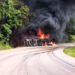 Militares do Corpo de Bombeiros usando mangueiras para apagar incêndio em carreta carregada com plástico na BR-364, com fumaça densa e área isolada na Serra de São Vicente