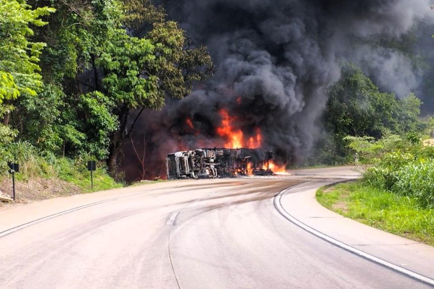 Militares do Corpo de Bombeiros usando mangueiras para apagar incêndio em carreta carregada com plástico na BR-364, com fumaça densa e área isolada na Serra de São Vicente