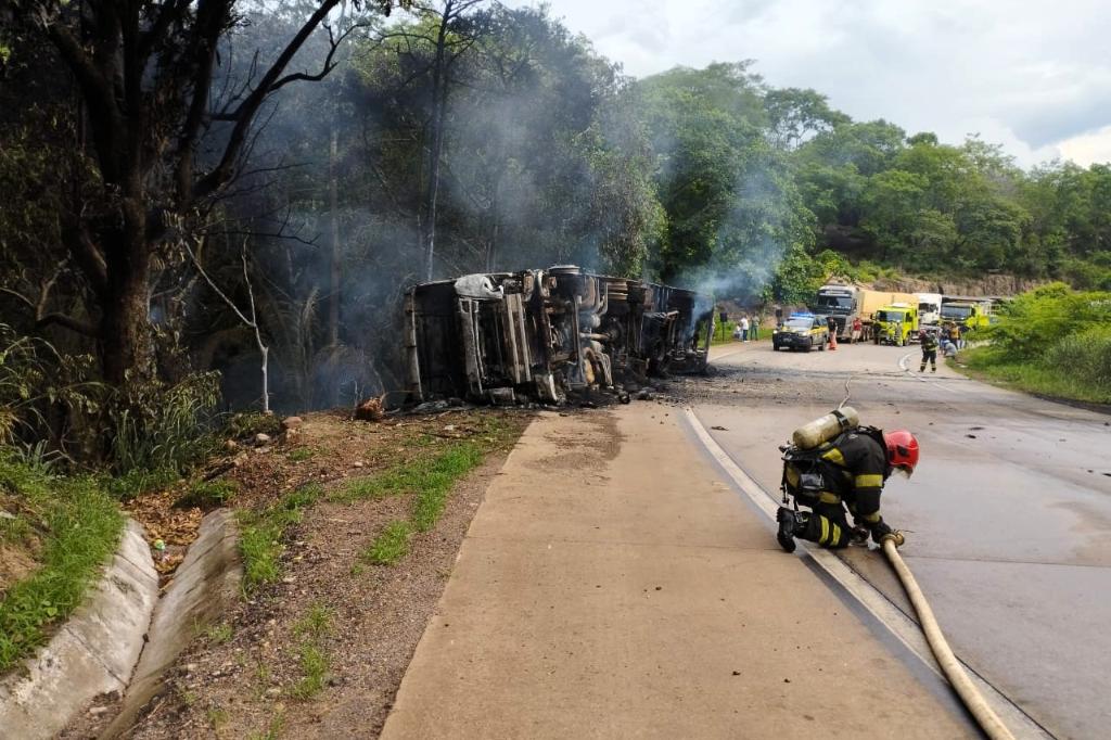 Militares do Corpo de Bombeiros usando mangueiras para apagar incêndio em carreta carregada com plástico na BR-364, com fumaça densa e área isolada na Serra de São Vicente