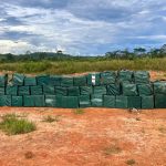 Policiais militares de Mato Grosso ao lado de dezenas de tabletes de apreensões de drogas em MT, em pátio de unidade policial