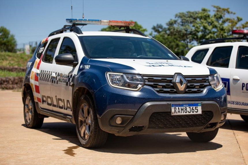 Imagem ilustrativa de uma viatura da Polícia Militar com as luzes de emergência ligadas em frente a um posto de combustíveis à noite.