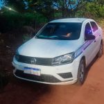 Carro branco estacionado em rua residencial com viatura da Polícia Militar ao fundo.