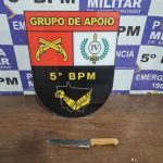 Imagem ilustrativa de uma faca apreendida sobre uma mesa policial com uma viatura ao fundo.